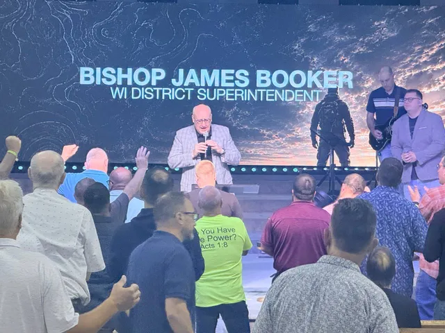Stage screen identifying Bishop James Booker as WI District Superintendent while he speaks.